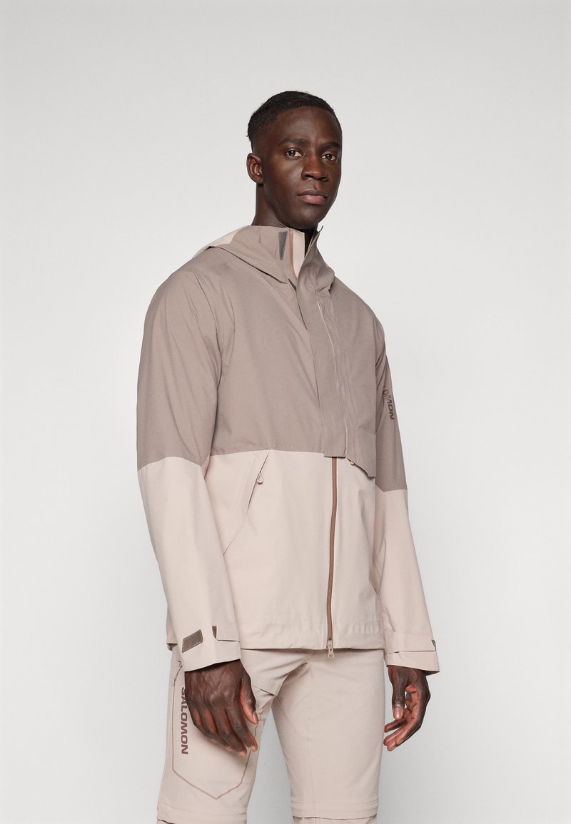 Lightweight, two-tone jacket in beige and taupe, featuring a high collar, zip closure, and zippered pockets. Made of smooth fabric.