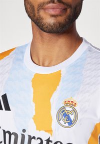 Maglia bianca e arancione con un motivo testurizzato, caratterizzata dal logo del Real Madrid e dal marchio Adidas in nero. Collo corto e rotondo.