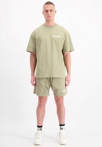 Olijfgroene katoenen t-shirt en bijpassende shorts met logodetail, relaxte pasvorm en zachte textuur, gecombineerd met witte sneakers en zwarte sokken.