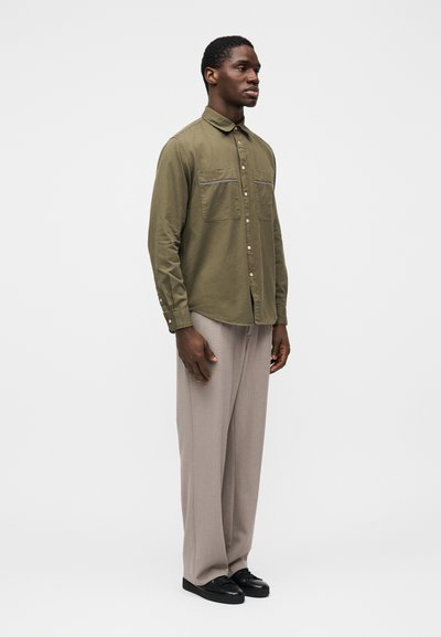 Πουκάμισο πράσινου χρώματος ελιάς με κουμπιά και δύο τσέπες στο στήθος, συνδυασμένο με φαρδιά παντελόνια ανοιχτού γκρι και μαύρα παπούτσια. Λείο ύφασμα, άνετη εφαρμογή.