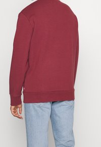 Sudadera burdeos de algodón, con cuello redondo, mangas largas, puños acanalados y dobladillo recto, llevada sobre unos jeans de mezclilla azul claro.
