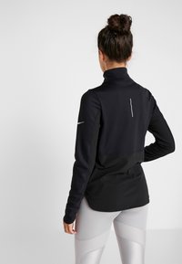 Sort sportsjakke med høj krave, glat tekstur og en diagonal søm på tværs af ryggen. Indeholder et lille reflekterende logo på ærmet.