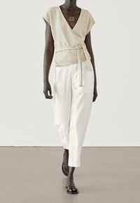 Beige wrap top with short sleeves and waist tie, paired with white, wide-legged trousers. Model wears black flip-flops, silver pendant necklace.