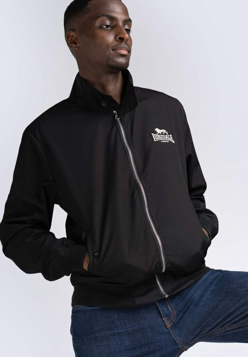 Chaqueta bomber ligera negra con cremallera frontal, cuello alto y puños acanalados. Cuenta con un logo bordado en el pecho. Corte casual.