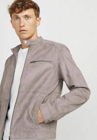 Giacca in suede grigio chiaro con colletto alto, taschino con zip sul petto e tasche laterali, caratterizzata da una texture liscia e linee pulite.