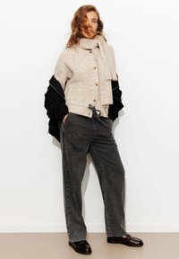 Cardigan en tricot beige clair avec des boutons, associé à un pantalon large gris foncé taille haute. Écharpe en laine et couche extérieure texturée noire. Chaussures marron.