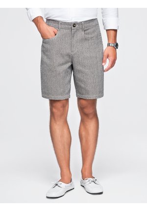 Homme portant un short gris texturé, une chemise blanche à manches longues, une montre-bracelet et des chaussures blanches décontractées, debout avec une main dans la poche.