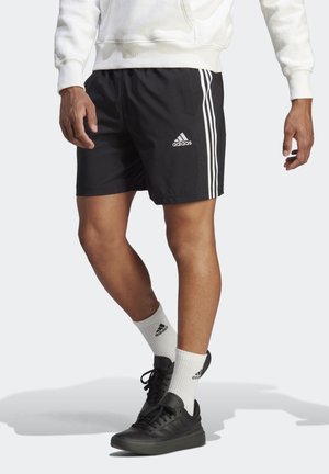 Mand iført sorte Adidas-shorts med hvide striber, hvide Adidas-strømper, sorte sneakers og en hvid sweatshirt, stående på en lyse farvet overflade.