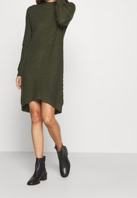 Abito maglione verde oliva a maglia con maniche lunghe, caratterizzato da un motivo a coste testurizzate e un orlo asimmetrico. Indossato con stivaletti neri alla caviglia.