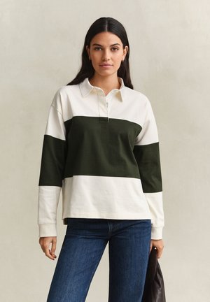 Femme aux longs cheveux foncés portant un t-shirt à manches longues blanc et vert foncé en blocs de couleur et un jean bleu, tenant une veste foncée.