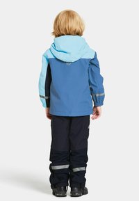 Barn med blont hår som bär en blå och svart vattentät jacka och byxor, vänd bortåt, stående mot en vit bakgrund.