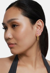 Des boucles d'oreilles en or en forme de créoles présentées sur l'oreille d'un modèle. Les boucles d'oreilles sont circulaires avec une finition lisse, réfléchissant la lumière sur un fond neutre.