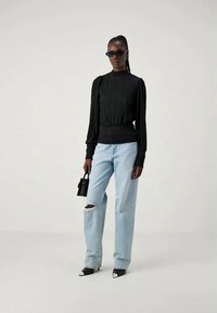Mulher vestindo uma blusa longa preta plissada com mangas, jeans rasgados azul claro, sapatos de salto pretos de bico, óculos de sol e segurando uma pequena bolsa preta.