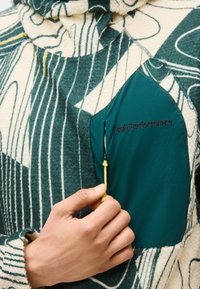 Ręka trzymająca żółty suwak na ciemnozielonej kieszeni wzorzystej polaru z logo Peak Performance.