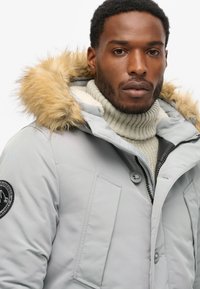 Chaqueta de plumas gris con capucha forrada de piel, con un logo en el pecho y detalles de botones. Un cuello de tortuga de lana por debajo añade textura.