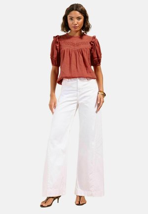 Frau steht und trägt eine rostfarbene Bluse mit Puffärmeln und Spitzen-Details, weiße, weit geschnittene Hosen, braune Sandalen mit Absatz und Armbänder.