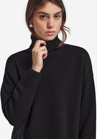 Sort turtleneck sweater med ribbet krave og manchetter; strikket tekstur; afslappet pasform. Modellen har en neutral udtryk, hånd nær hagen.