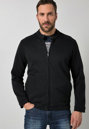 Mann mit Bart, der eine schwarze Zip-Up-Jacke über einem gemusterten dunklen Hemd und blauen Jeans trägt, steht mit einer Hand in der Tasche vor einem einfarbigen Hintergrund.