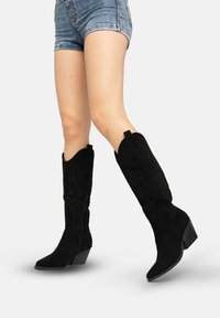 Schwarze Cowboy-Stiefel aus Wildleder mit spitzen Zehen, einem geschwungenen Schaft und dekorativen Nähten. Sie verfügen über einen geschichteten Absatz und einen seitlichen Reißverschluss.