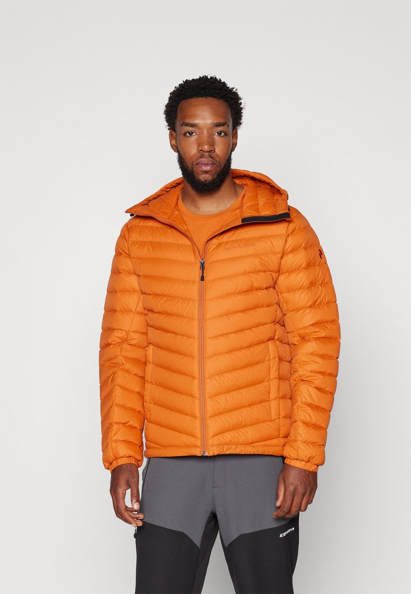 Orange pufferjakke med horisontal quiltet design, frontlynlås og justerbar hætte. Bæret over en matching orange skjorte, parret med grå bukser.