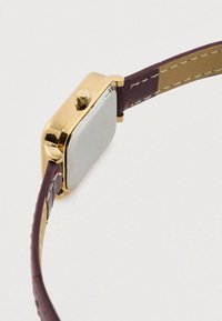 Stačiakampis geltono aukso atspalvio laikrodžio korpusas su lygiu sidabriniu paviršiumi, derinamas su tamsiai violetinės odos dirželiu su siuvinėjimo akcentais.