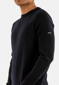 Maglione lavorato a maglia blu navy con scollo rotondo, maniche lunghe e una piccola etichetta sul braccio sinistro. Texture liscia, design aderente.