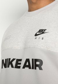 Nära bild av en ljusgrå Nike Air sweatshirt som bärs av en person, med svarta Nike-logotyper och text på bröstområdet.
