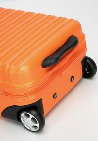 Valigia rigida arancione con texture a righe, manico nero e angoli rinforzati. Dotata di quattro ruote nere per la mobilità.