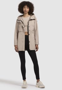 Beige waterdichte jas met capuchon, ritssluiting aan de voorkant en een dierenprintvoering, gecombineerd met een aansluitend topje en zwarte leggings.