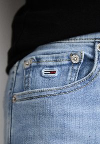 Jeans de mezclilla azul claro con una pequeña etiqueta de logo que presenta colores rojo, blanco y azul en el bolsillo trasero de la cintura. Detalles de botón de metal visibles.