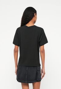 Sieviete ar gariem viļņainiem matiem valkā vienkāršu melnu īss piedurkņu t-kreklu un melnu džinsa mini svārku ar kabatu atloki, skatoties prom.