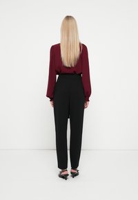 Long-sleeve burgundy blouse with puffed shoulders paired with high-waisted black trousers. Model wears black heeled sandals.