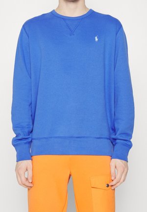 Person iført en blå langærmet sweatshirt med et lille hvidt logo og lyse orange bukser med en sidelomme og knapklap.