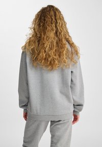 Persona con lunghi capelli ricci biondi che indossa una felpa grigia larga e pantaloni felpati grigi abbinati, girata di spalle su uno sfondo semplice.