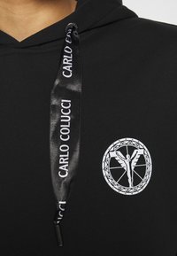 Sweat à capuche noir avec cordon de serrage en satin noir imprimé "Carlo Colucci" et un logo circulaire blanc avec une figure ailée sur la poitrine.