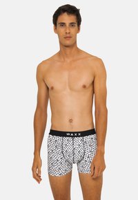 Boxers pour hommes avec une ceinture noire portant l'inscription "WAXX". Le corps est blanc avec un motif noir répété de petites formes. Tissu doux au toucher.