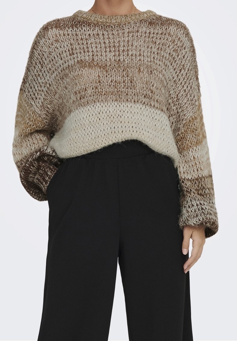 Femme portant un pull en maille ample aux tons marron et crème avec un pantalon noir taille haute, main dans la poche sur un fond uni.