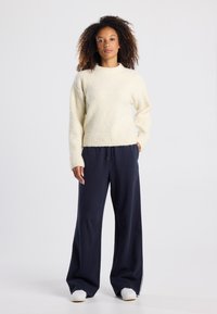 Maglione morbido color crema con scollatura rotonda e maniche lunghe, abbinato a pantaloni della tuta a gamba larga di colore blu navy e sneakers bianche. Tessuto liscio.