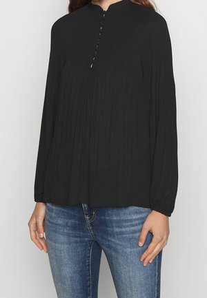 Nainen pukeutuneena pitkähihainen musta paita, jossa etuosassa nappidetaljit, yhdistettynä sinisiin istuviin farkkuihin vaaleaa yksinkertaista taustaa vasten.