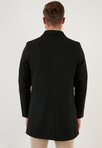 Cappotto nero in misto lana con taglio sartoriale, maniche lunghe e un'unica spacco posteriore. Presenta polsini con bottoni e una texture liscia.