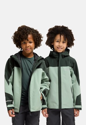 Zwei Kinder tragen hellgrüne Jacken mit dunkelgrünen Akzenten, die einen Reißverschluss, eine Kapuze und reflektierende Details aufweisen. Die Jacken haben ein geometrisches Design.