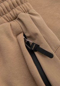 Gros plan sur un pantalon beige à taille élastique avec une fermeture éclair noire et un curseur en caoutchouc portant la mention « OMBRE » sur la poche latérale.
