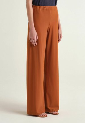 Persona che indossa pantaloni a gamba larga color ruggine con vita elastica e sandali bassi marroni, in piedi davanti a uno sfondo neutro.