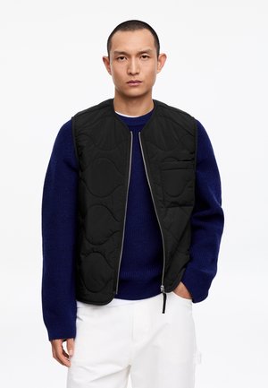 Mann trägt eine schwarze gesteppte Weste über einem blauen Pullover und weißen Hosen, steht mit einer Hand in der Tasche vor einem einfarbigen weißen Hintergrund.