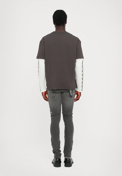 Grijze T-shirt met korte mouwen laag over een wit T-shirt met lange mouwen, gecombineerd met grijze skinny jeans en zwarte schoenen, gezien van achteren.
