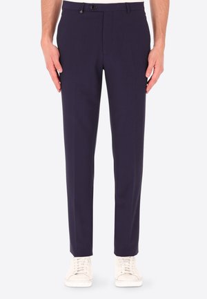 Pantaloni slim fit da uomo blu navy con chiusura con bottone e zip, indossati con sneakers bianche con lacci, mostrati dalla vita ai piedi.