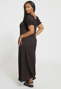 Kvinna som poserar i profil i en svart maxiklänning med rosa och vita blommönster, puffärmar och svarta sandaler med spännen.