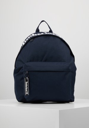 Marineblauer Timberland-Rucksack mit vorderer Reißverschlusstasche, Tragegriff oben und sichtbarem Logo am Reißverschluss und am oberen Rand, auf einem weißen Podest platziert.