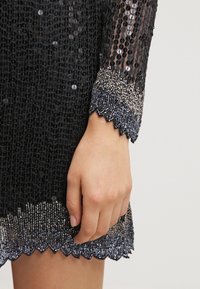 Robe noire à sequins avec un tissu texturé, un ourlet festonné et des perles brillantes ornant les poignets. Main visible au bas de la photo.