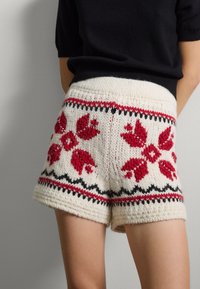 Pantalones cortos de punto en color crema con patrones florales rojos y acentos negros, que cuentan con un acabado texturizado y dobladillo doblado, adecuados para vestir de forma casual.